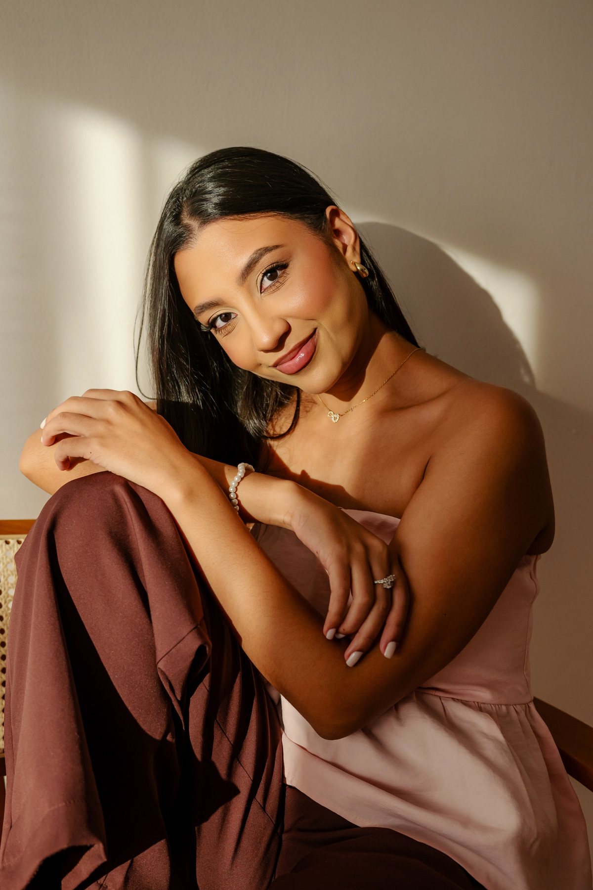 Giovanna sorrindo, sentada em uma cadeira de palhinha, com luz natural na parede.