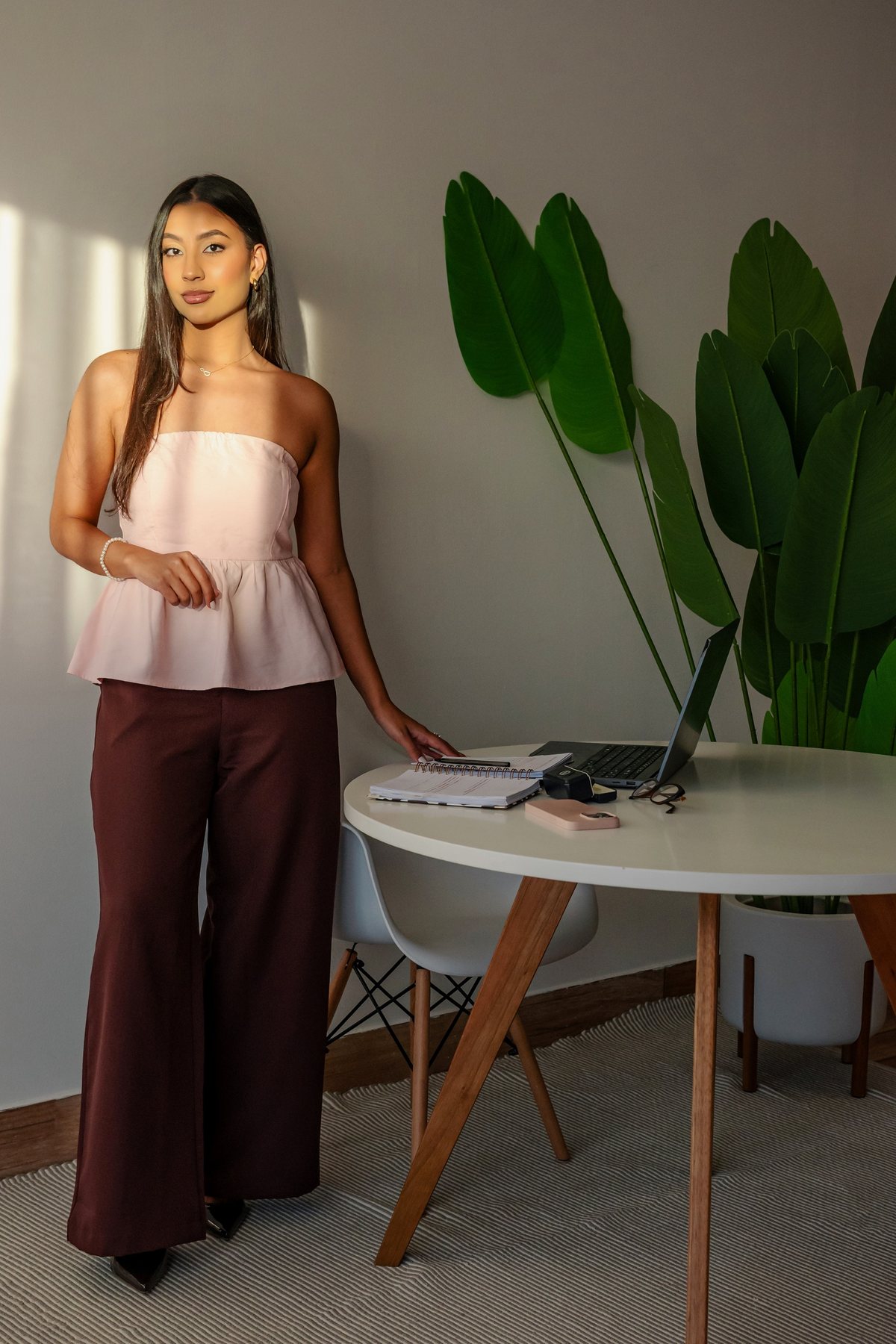Giovanna Alves em pé, ao lado de uma mesa branca com caderno e notebook, em luz natural de fim de tarde.