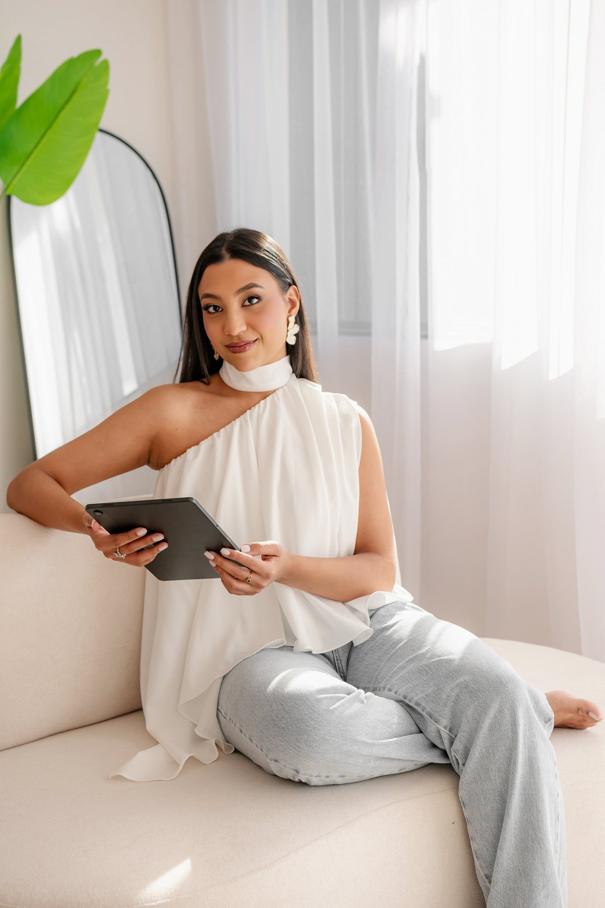 Giovanna sentada em um sofá claro, segurando um tablet, ao lado de uma janela com cortina branca.